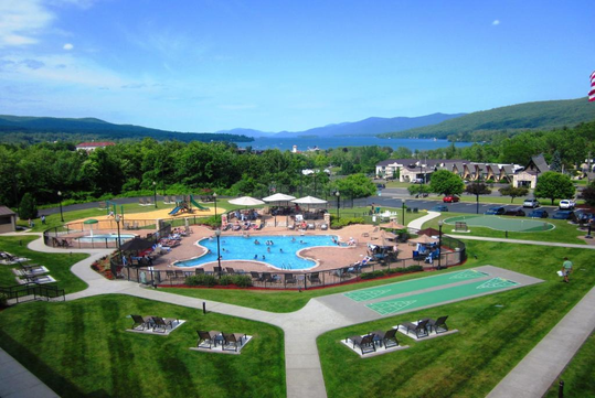 Aerial view of a resort with a central swimming pool, surrounded by green lawns, a playground, shuffleboard courts, and nearby houses, with a scenic lake and hills in the background.