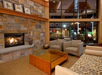 Cozy living room with a stone fireplace, glass coffee table, gray chairs, and a patterned sofa. Large windows reflect interior lights and wooden ceiling accents.