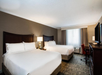 Hotel room with two double beds, a nightstand with a lamp, a window with curtains, a desk with a chair, and a TV on a dresser.
