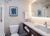 A modern bathroom with a lit mirror, sink, and toiletries on a white countertop. There's a towel rack above a toilet and a framed abstract artwork on the wall.