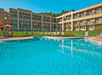 A curved outdoor pool with lounge chairs and umbrellas is in front of a multi-story building with balconies.