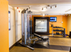 A gym with exercise equipment including a weight machine, a bench, and a rack of dumbbells. A wall-mounted TV is on, and a large mirror is visible.