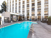 Outdoor hotel pool surrounded by lounge chairs and a tall building with multiple balconies. The pool area has safety signs and railings.