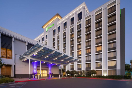 A modern hotel building with multiple floors, featuring a glass entrance canopy and illuminated signage. The scene is captured during the evening with exterior lights on.
