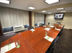 A conference room with a long wooden table, empty chairs, and a TV on the wall. Notepads and glasses are placed on the table. The room has carpeted flooring and curtained windows.