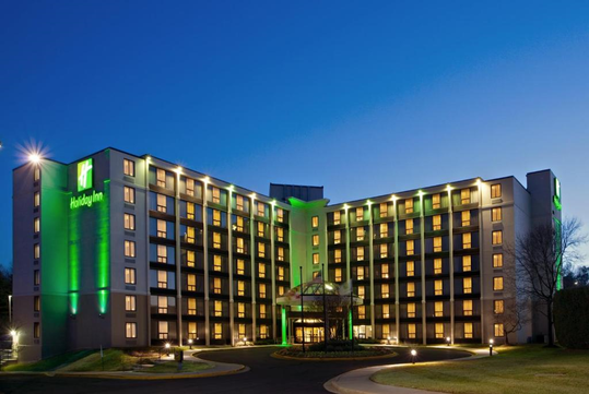 A multi-story Holiday Inn hotel illuminated at night, featuring green and white signage, with a lit entrance and neatly maintained landscaping.