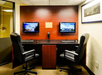 Two office chairs facing a wooden desk with two computer monitors on the wall, displaying images of a hotel room and exterior. A black handset is between the screens.