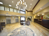 Hotel lobby with marble flooring, a circular rug, a chandelier, elevators on the left, and a reception desk on the right. Seating area with chairs and a table in the center.