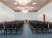 An empty conference room with rows of black chairs facing a wooden podium. Chandeliers hang from the ceiling, and the room has blue-patterned carpet.