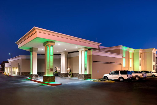 Hotel entrance at dusk with illuminated pillars and green lighting accents. Four parked vehicles are visible in front.