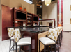 Modern bar interior with a granite countertop, checkered upholstered chairs, and shelves stocked with liquor bottles. Two TV screens are mounted on the back wall.