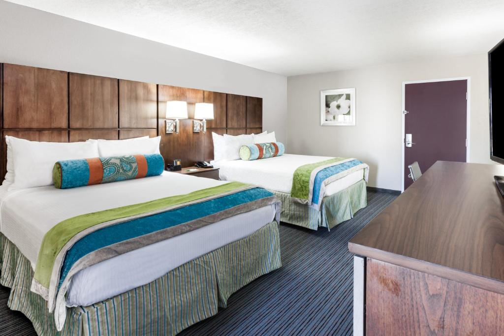 Hotel room with two double beds, colorful pillows, a wooden headboard, wall art, a lamp, a flat-screen TV on a wooden dresser, and a closed purple door.