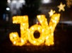 The image shows the word "Joy" spelled out in bright, out-of-focus yellow lights with star shapes in the background.