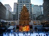 People skate on an ice rink in front of a large, decorated Christmas tree at Rockefeller Center, surrounded by tall buildings and flags.