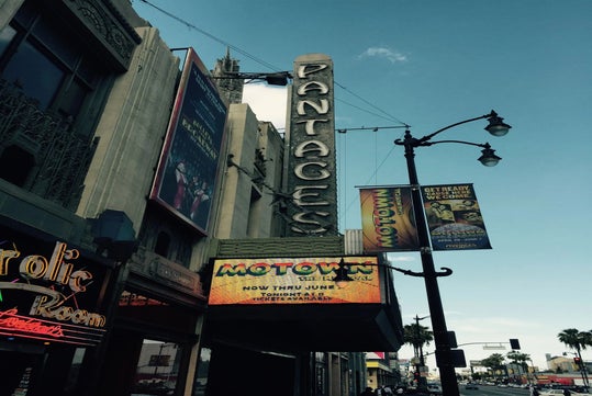 Vertical sign for Pantages Theatre and marquee advertising “Motown the Musical,” with surrounding street and palm trees visible under a clear sky.