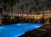 Outdoor hotel pool at night illuminated with blue lights, surrounded by lounge chairs, palm trees, and buildings with string lights.