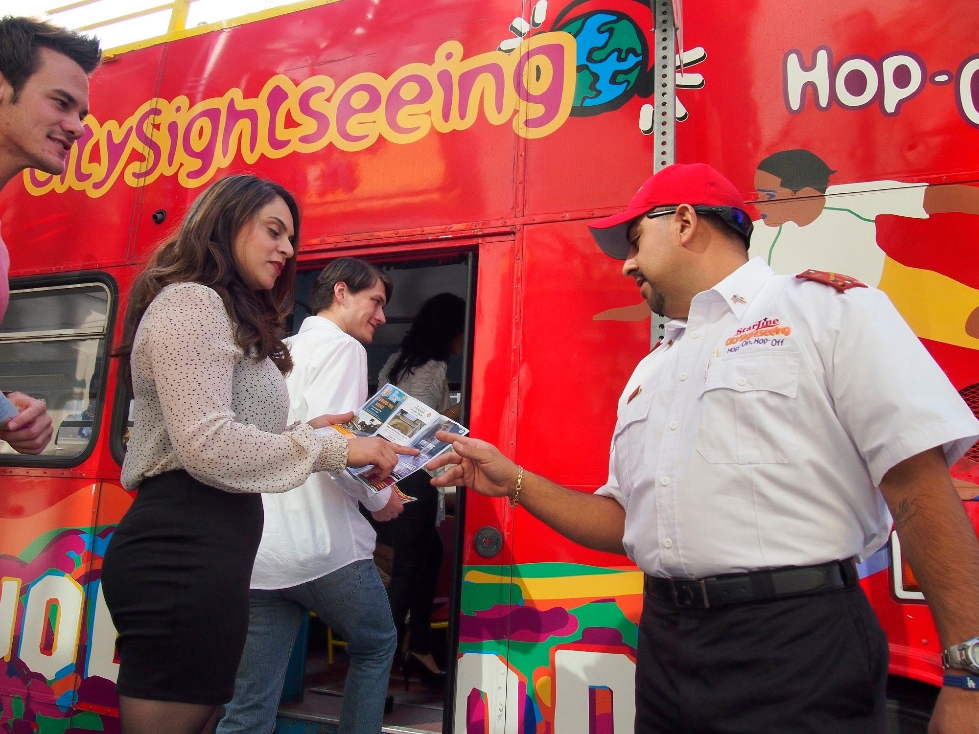 People boarding a red sightseeing bus. A uniformed staff member hands out brochures to a woman wearing a black and white outfit. The bus has colorful designs and text reading "sightseeing.
