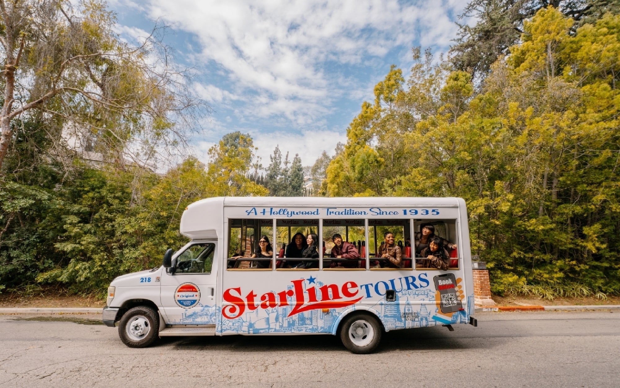 A Starline Tours bus with passengers drives on a road surrounded by trees under a partly cloudy sky.