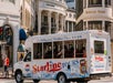 A StarLine Tours bus drives through a city intersection, passing upscale shops including Dolce & Gabbana and Stefano Ricci. Passengers are seated inside the bus.