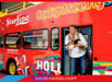 A man holding a small dog stands in the doorway of a red double-decker CitySightseeing bus decorated with Hollywood-themed artwork.