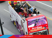A city sightseeing bus with "Speak Now" and "Taylor Swift" on the side carries musicians performing on the upper deck as it travels down a street.