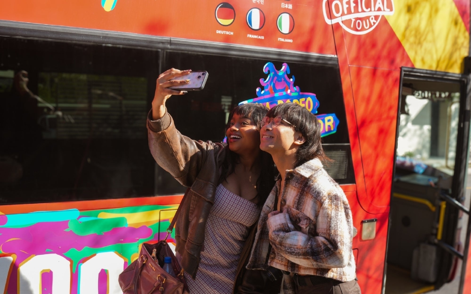 Two people stand in front of a red double-decker tour bus, smiling and taking a selfie together in bright daylight.
