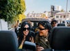 A group of young people ride in a convertible car on a sunny day, smiling, wearing sunglasses, and using their phones.