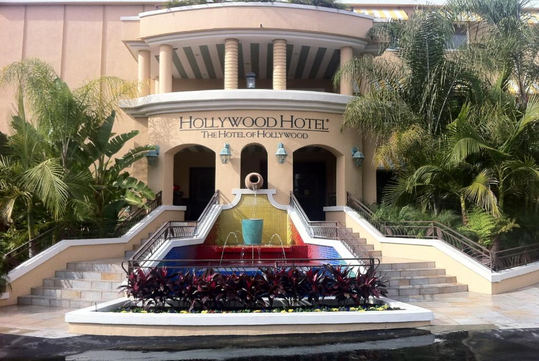 Entrance of Hollywood Hotel with a grand staircase, decorative fountain, and lush palm trees in the foreground.