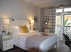 A cozy bedroom with a double bed, white bedding, a yellow decorative pillow, lamps, a hat, and a book on the bed. There are curtains and framed pictures on the walls.