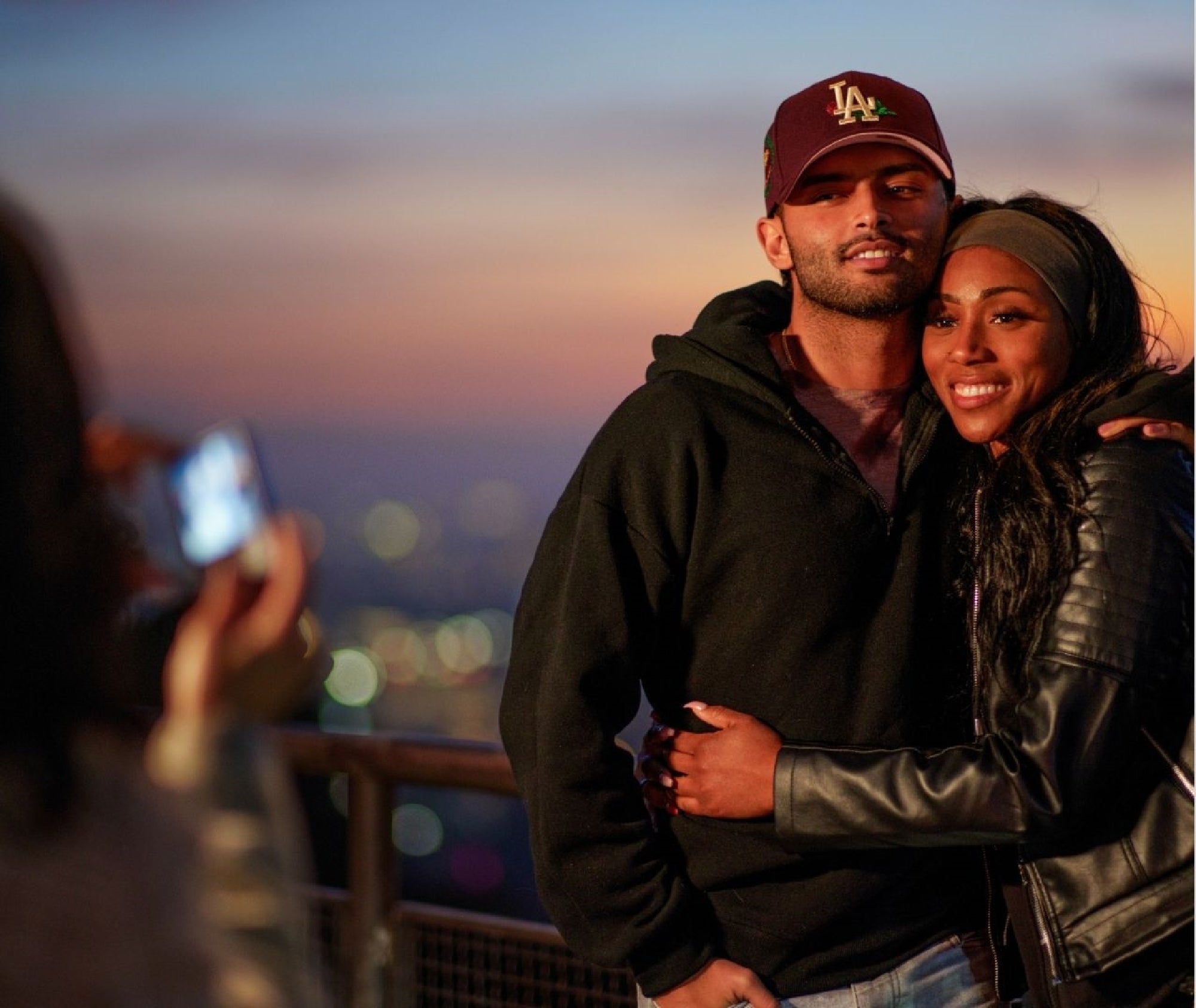 A man and woman stand close together, smiling as they pose for a photo at sunset with a blurred cityscape in the background.