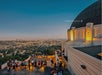 Griffith Observatory at sunset, overlooking downtown Los Angeles with visitors gathered on the outdoor terrace.