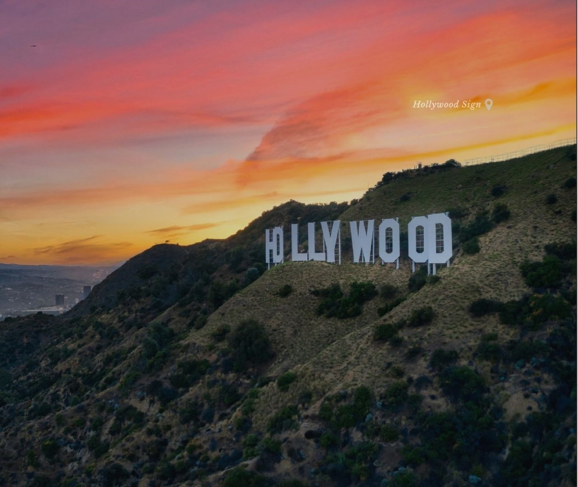 The Hollywood Sign stands on a hillside at sunset, with a colorful sky in the background and scattered shrubs on the slope.
