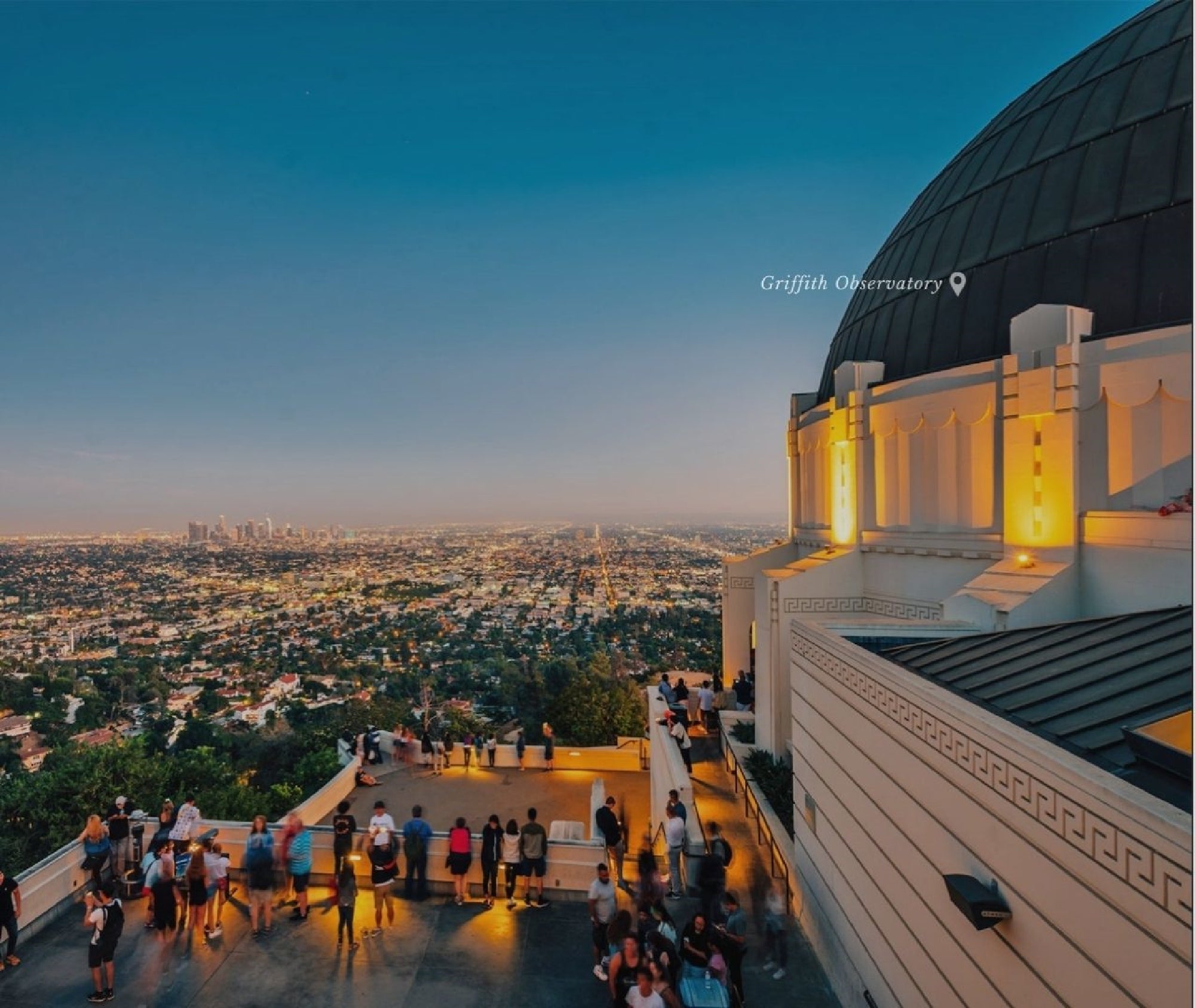 A crowd gathers on the terrace of Griffith Observatory at sunset, overlooking the cityscape of Los Angeles under a clear sky.