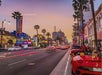 A view of Hollywood Boulevard at sunset with palm trees, neon signs, cars driving by, and a red convertible parked on the street.