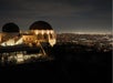 Griffith Observatory at night, illuminated against the dark sky, overlooks the city lights of Los Angeles in the background.