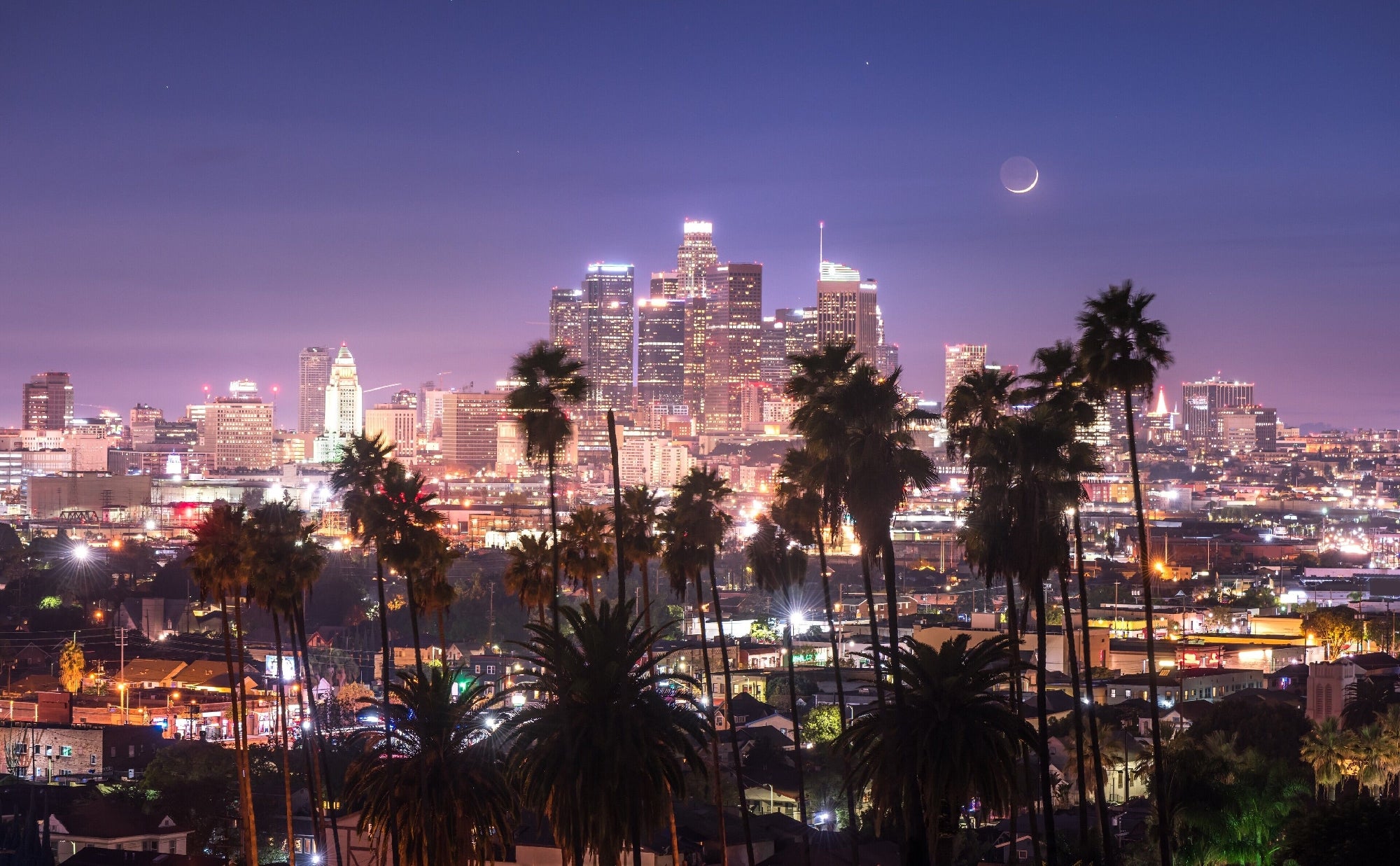 Downtown Los Angeles skyline at dusk with illuminated buildings, palm trees in the foreground, and a crescent moon in the sky.