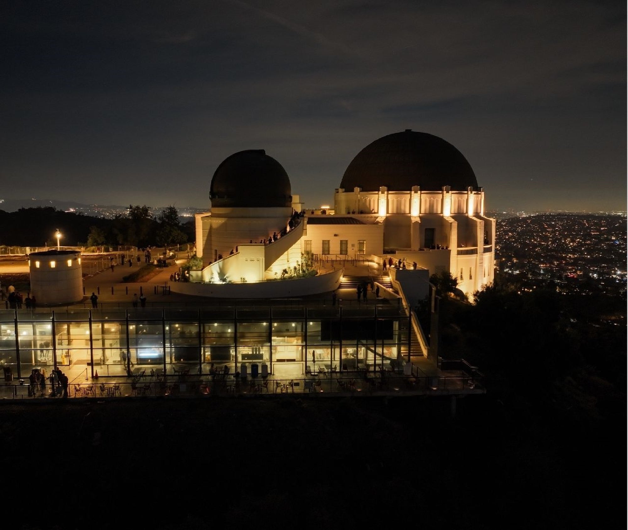 Griffith Observatory at night, illuminated with city lights visible in the background and visitors walking around the building and terraces.