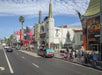 Busy Hollywood street with pedestrians, a colorful van, and iconic theaters. Buildings are adorned with signs and posters. Sky is partly cloudy.