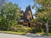 Whimsical house with a steep, pointed roof, surrounded by trees and greenery, located at a street corner under a clear blue sky.