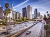 City skyline with tall buildings and a busy highway in the foreground, bordered by palm trees under a clear sky.