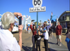 A group poses for a photo in front of the "Route 66 End of the Trail" sign on a boardwalk, while another person takes their picture.