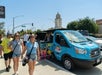 People walk near a parked blue tour van on a sunny street. A historic building is visible in the background.