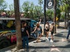 People standing near a parked, colorful tour bus on a sunny street lined with trees. A woman poses energetically in front of the bus, while others chat or wait nearby.