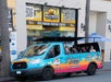 A colorful tour van labeled "Hollywood Bus Tours" is parked on the street in front of the Hollywood Visitor Center, where people are seated inside the open-air vehicle.