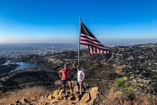 Two men in shorts and tee shirts standing next to an American Flag blowing in the wind on the Hollywood Sign Hike.