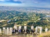 View looking down on a group of hikers standing behind the Hollywood sign with Los Angeles in the distance.