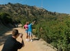 Two people pose for a photo with the Hollywood sign in the background on a sunny day, while another person takes their picture on a dirt path.