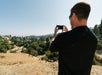 Person in a black shirt and sunglasses taking a photo with a smartphone, overlooking a dry landscape with trees and hills under a clear sky.