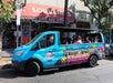 Blue open-top van labeled "Hollywood Bus Tours" parked on a street with a souvenir shop in the background. Passengers are seated inside, and the driver is waving.