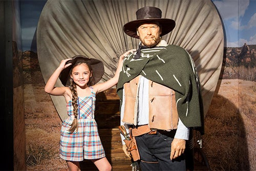 A young girl in a plaid dress poses next to a cowboy wax figure in front of a covered wagon backdrop.